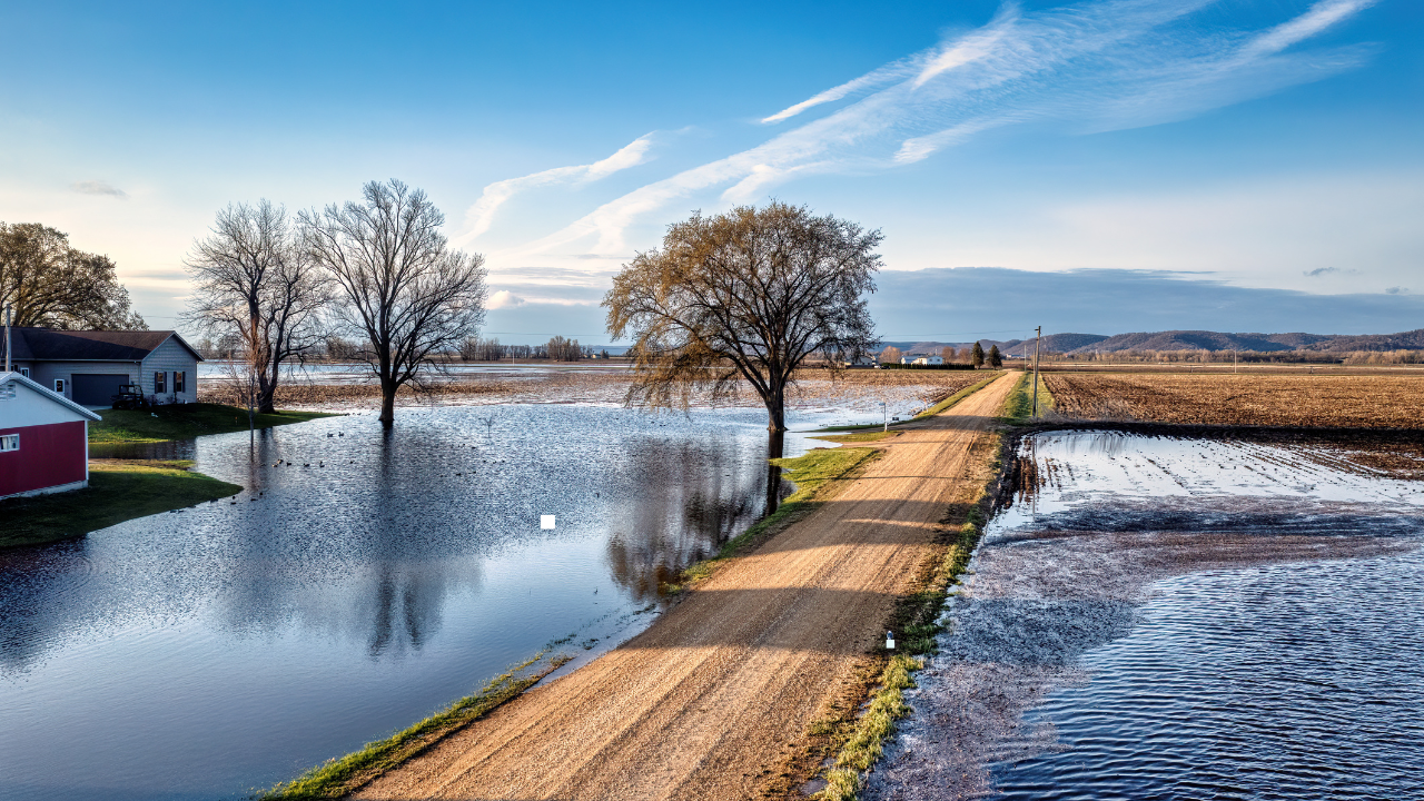 Flooded rural property with water covering fields and surrounding a home near a dirt road, illustrating flood risk and damage