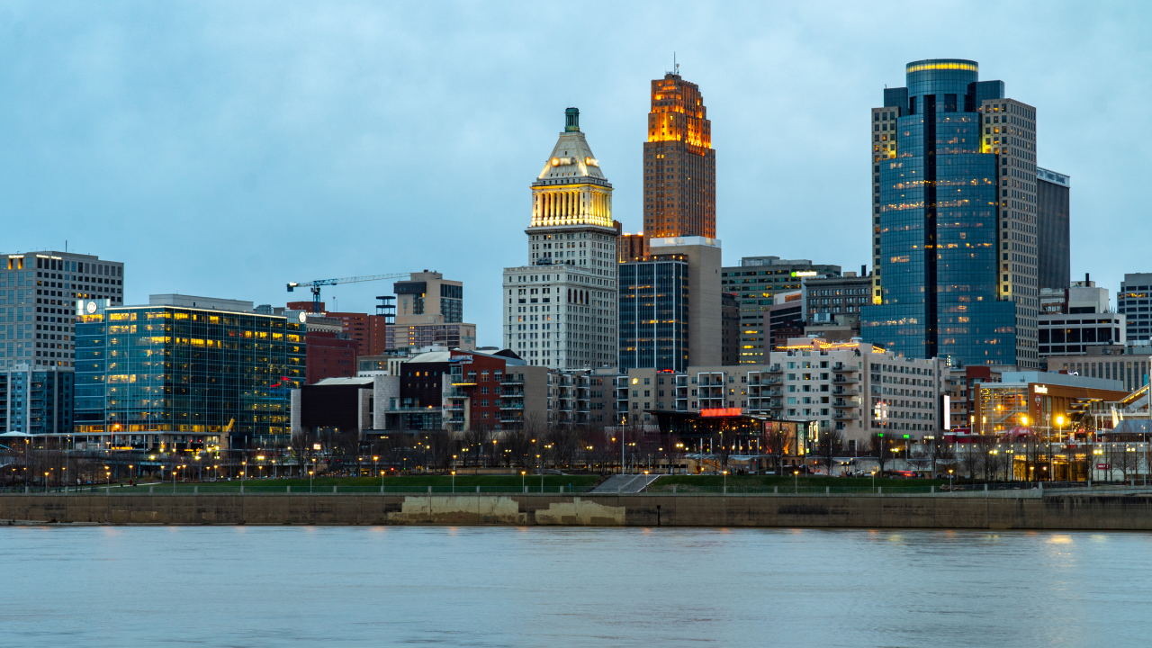 City skyline of Cincinnati at dusk, featuring illuminated downtown buildings along the Ohio River waterfront.