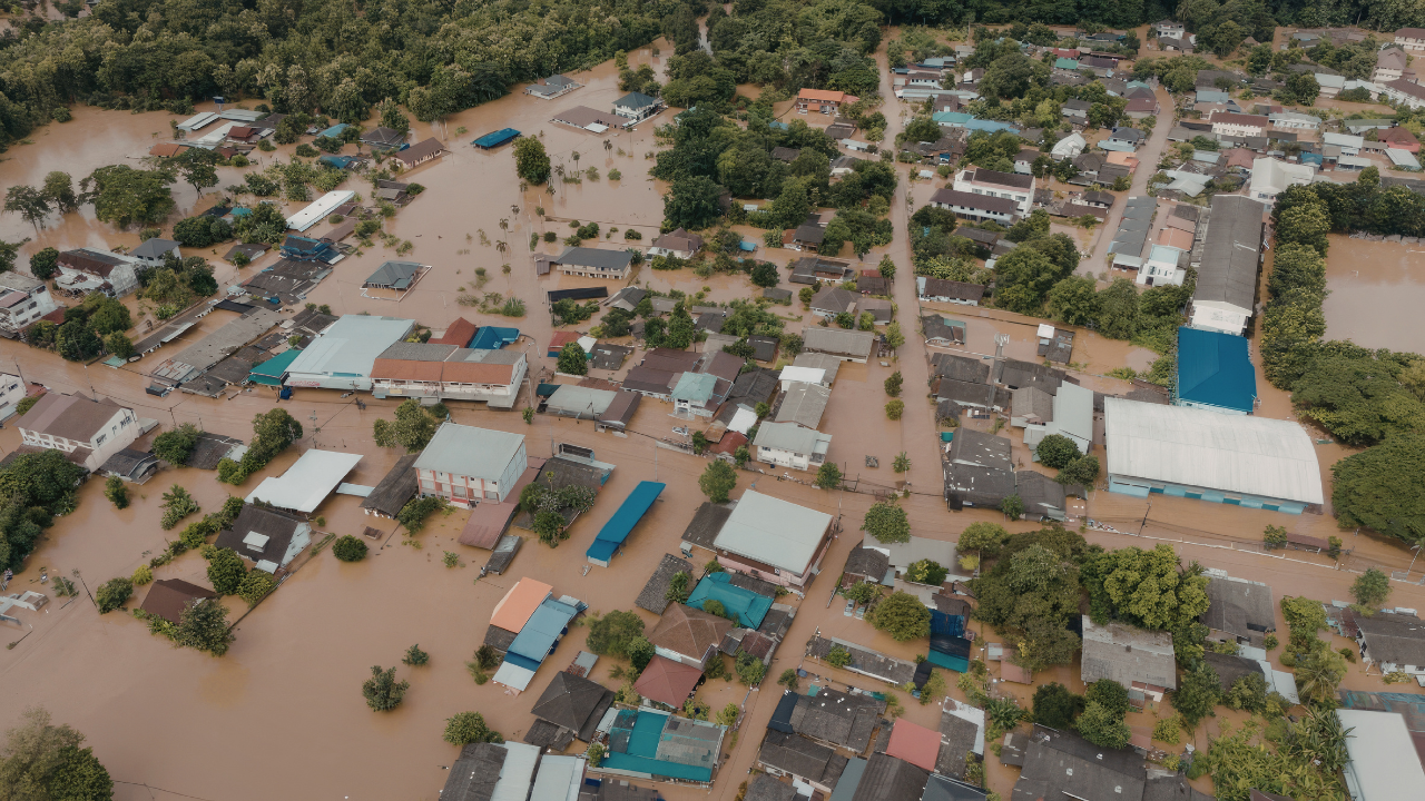 Aerial view of a flooded town with homes, buildings, and streets submerged in muddy floodwater surrounded by trees and residential neighborhoods.