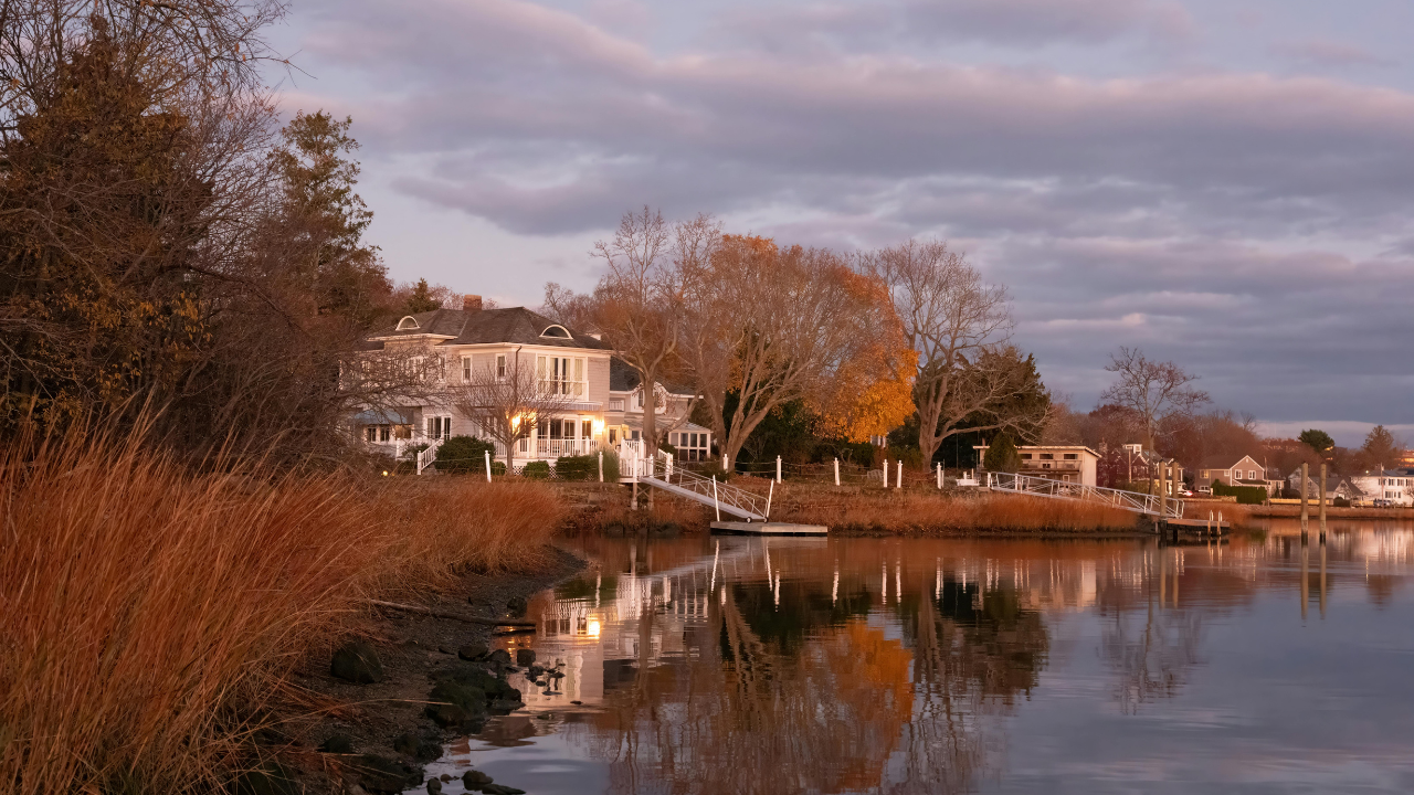 Waterfront homes along a calm Connecticut shoreline at sunset with reflections on the water, autumn trees, and cloudy evening skies.