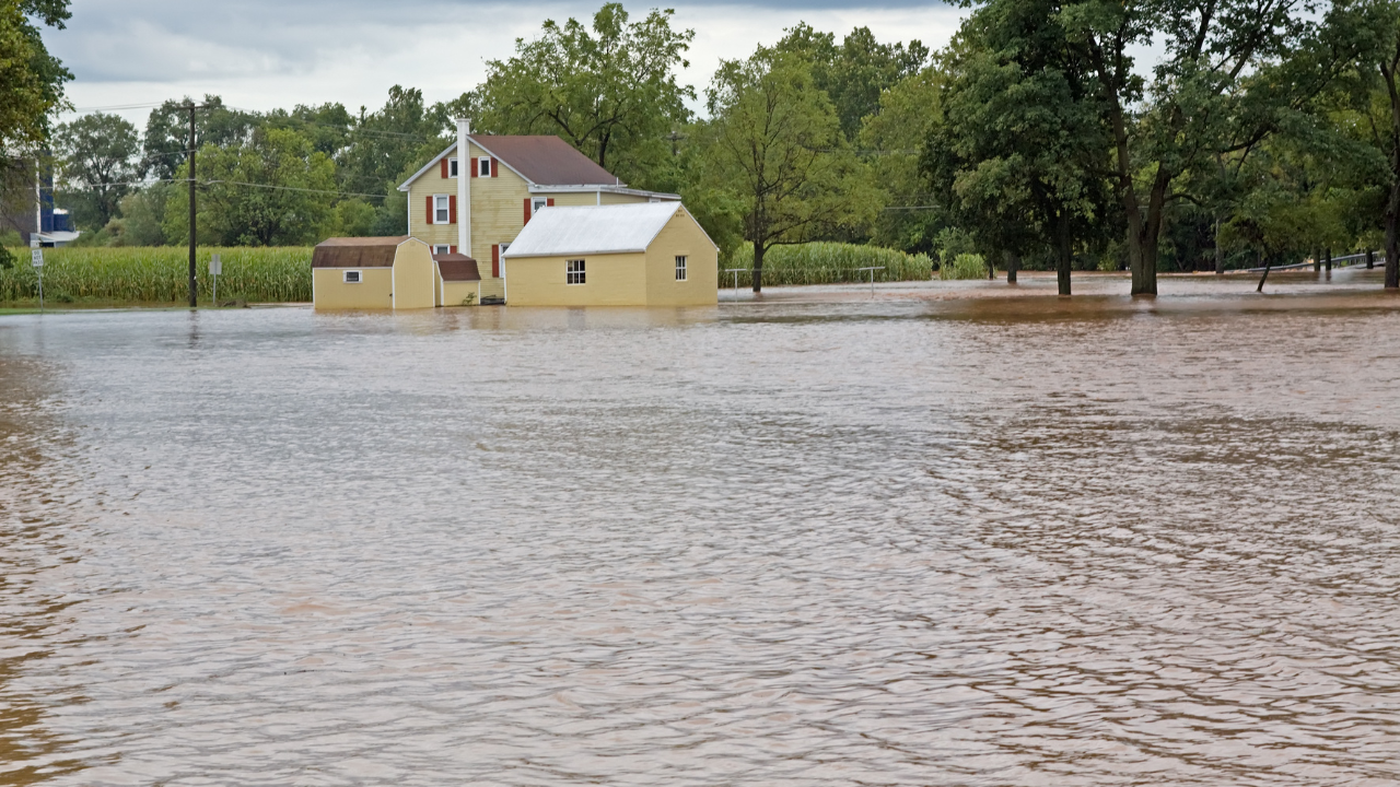 Flooded rural home surrounded by high water after severe flooding, with trees, outbuildings, and overcast skies in the background.