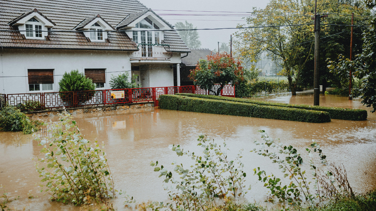 Flooded residential property with standing water surrounding a suburban home, trees, and yard after heavy rainfall and severe flooding.