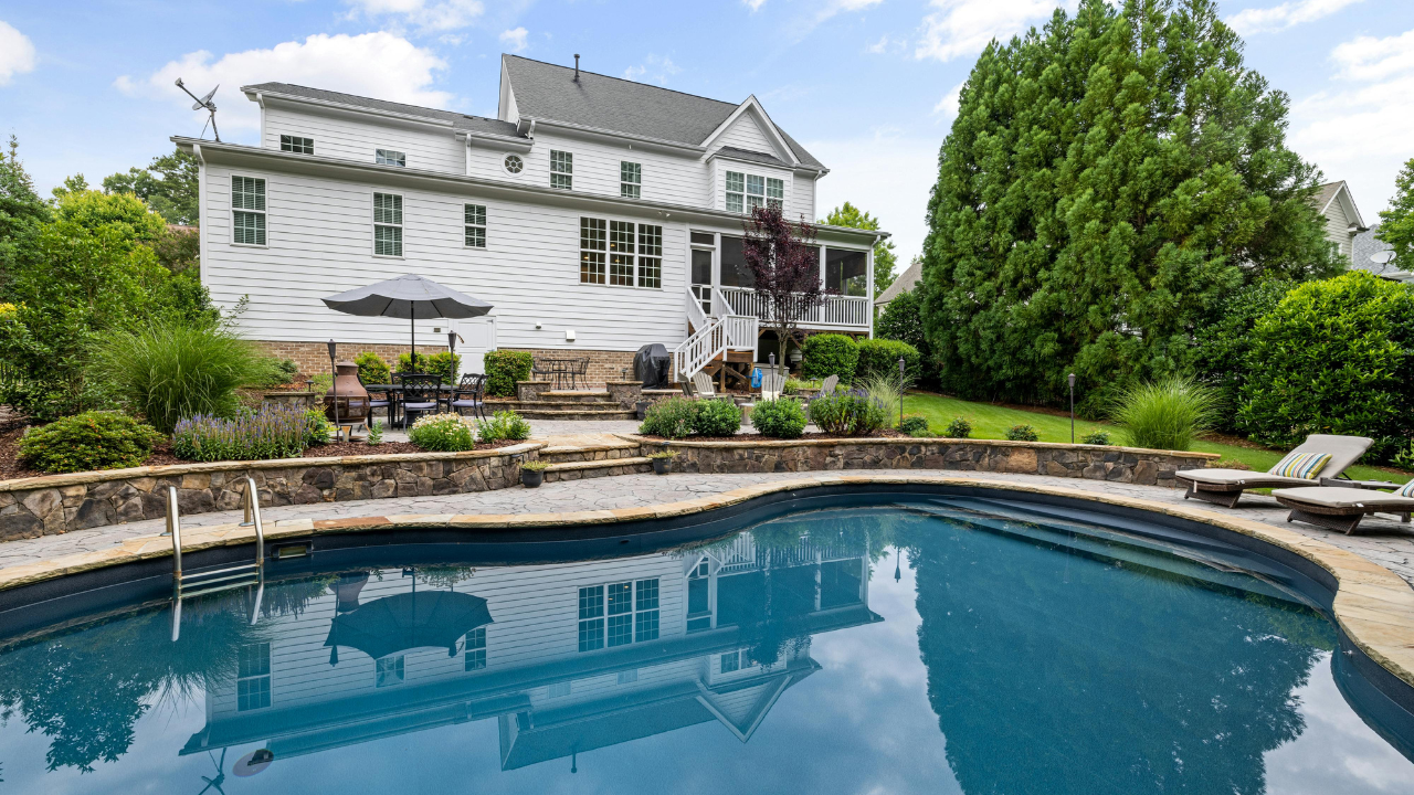 Backyard swimming pool behind a large residential home with a stone patio, outdoor seating area, landscaped yard, and clear blue water on a sunny day.