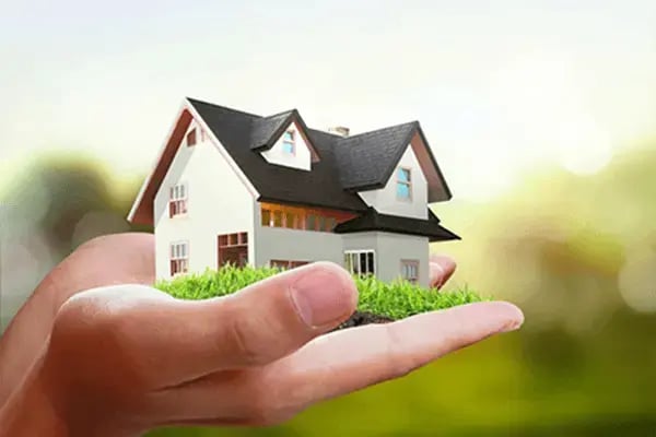 A miniature house sitting on a person's open palm with a bright green nature background