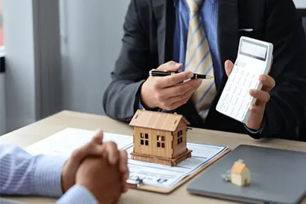 A loan officer pointing with a pen during a mortgage meeting with a wooden model house on the desk