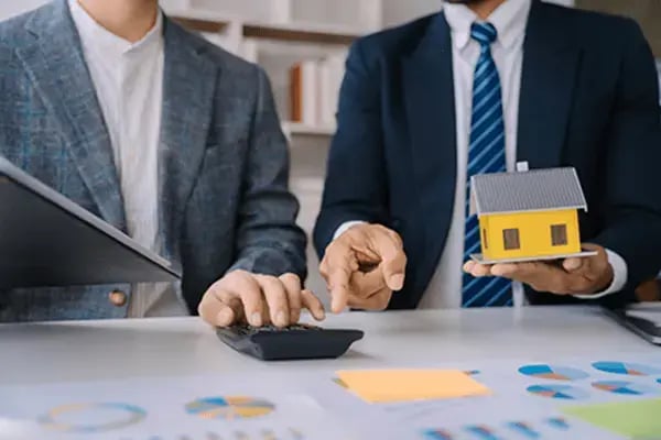 Two businessmen reviewing financial charts on a desk while holding a small yellow model home